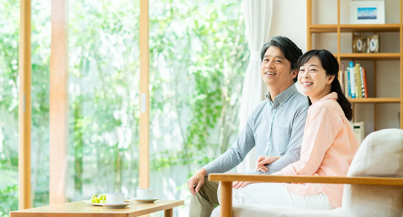 明るいリビングで、仲良くソファに座る中年夫婦。窓の外には緑が広がり、リラックスした雰囲気が漂う。